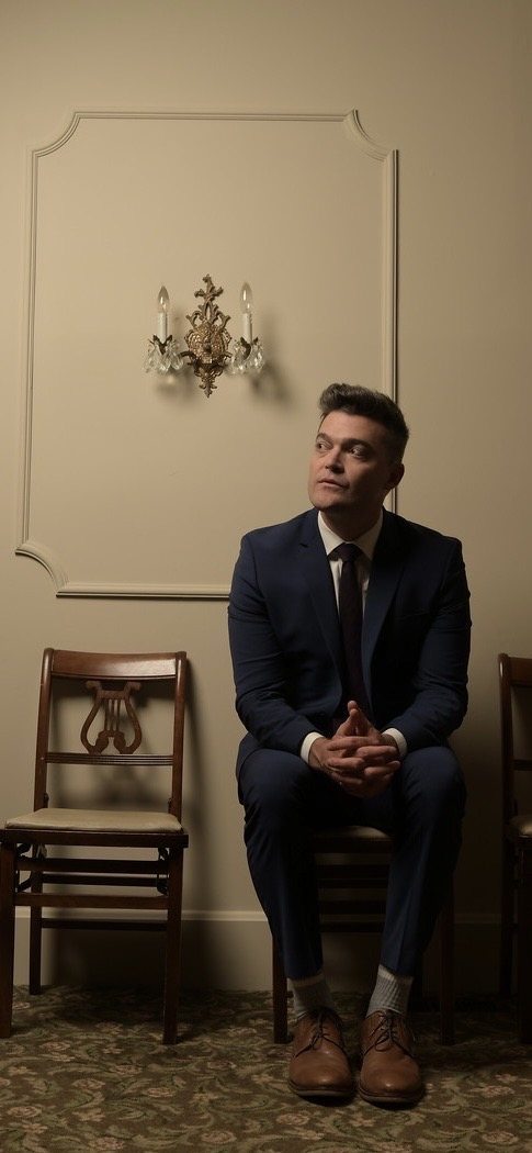 Nathan Morris seated upright in a dark suit in a funeral parlor, rows of chairs and a wall sconce behind him.