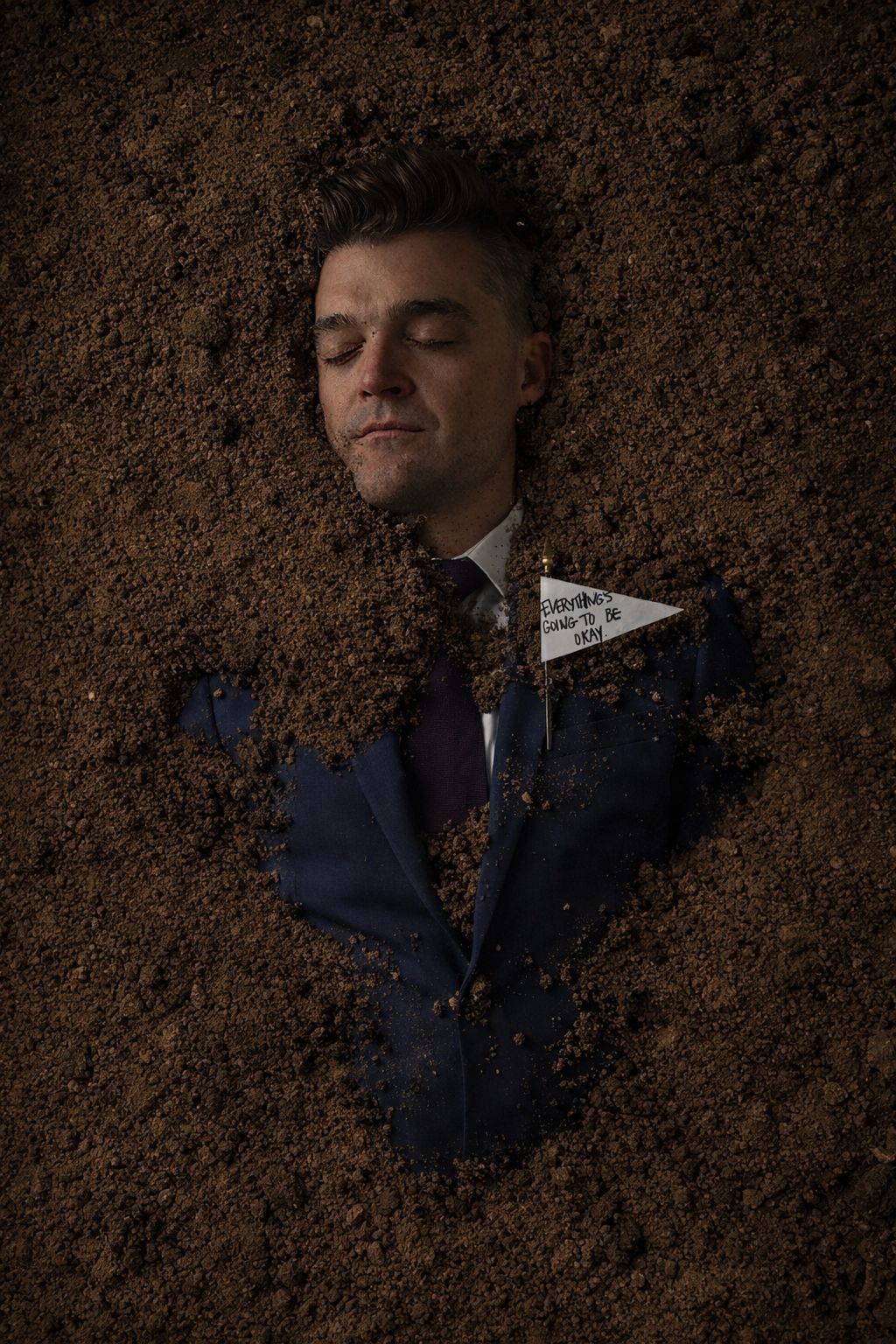 Nathan Morris buried in soil in a navy suit, a small flag reading Everything's going to be okay pinned to his lapel.