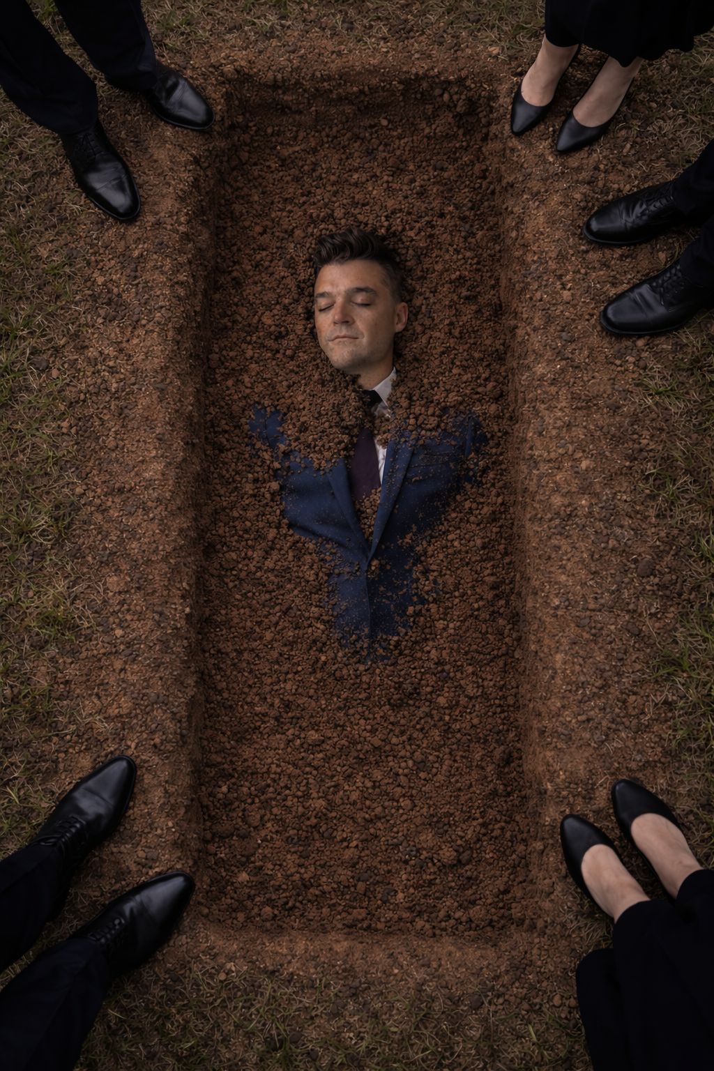 Overhead view of Nathan Morris buried to the chest in a grave, with mourners' black shoes around the edge.