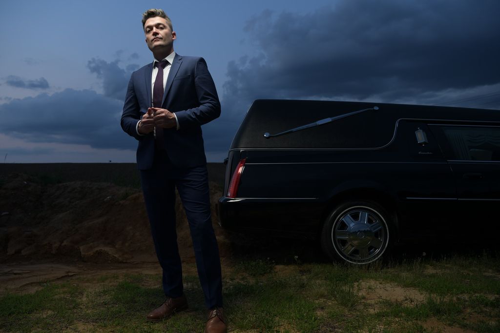 Nathan Morris in a navy suit standing beside a hearse at dusk.