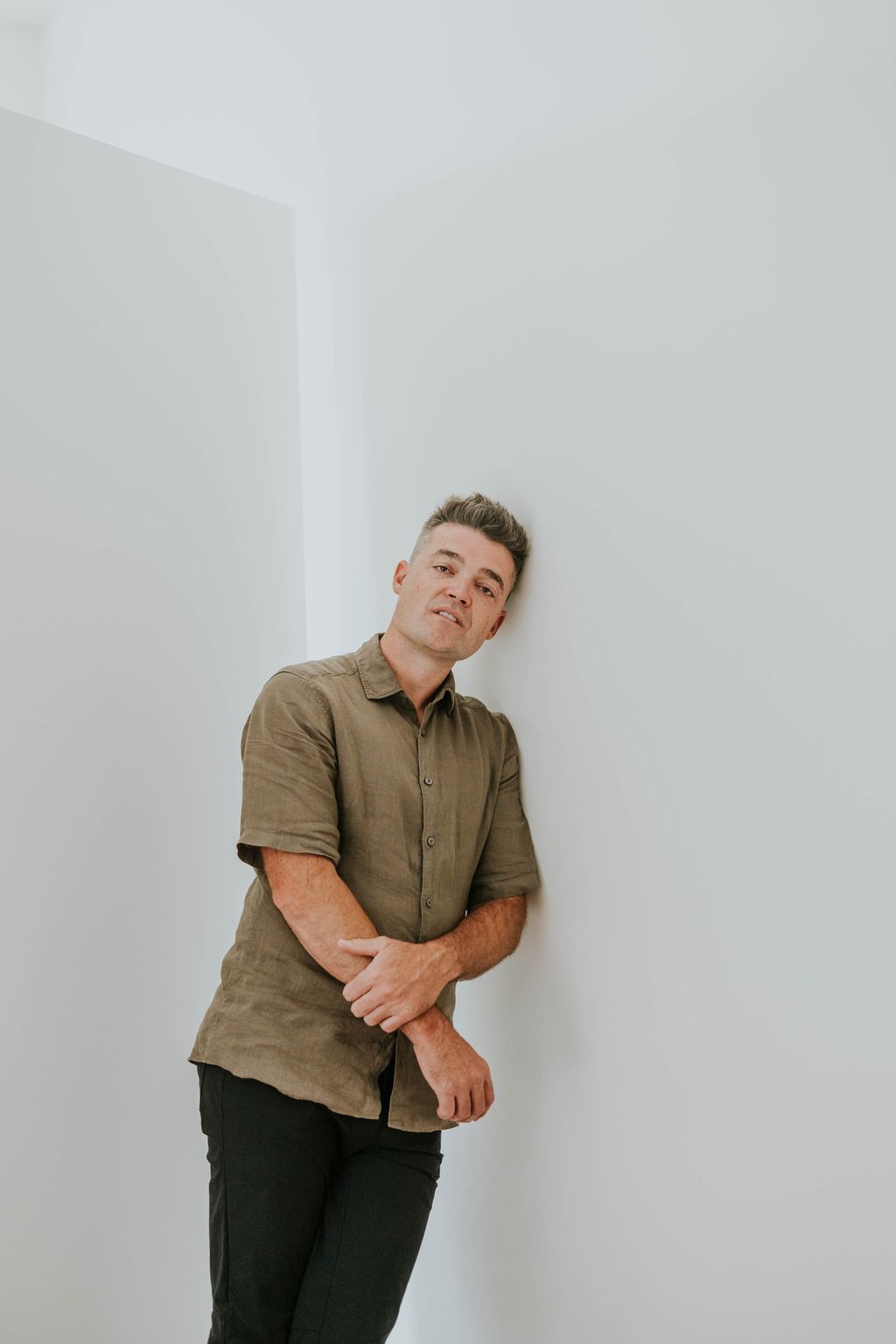 Nathan Morris in an olive linen shirt leaning his head against a white wall.