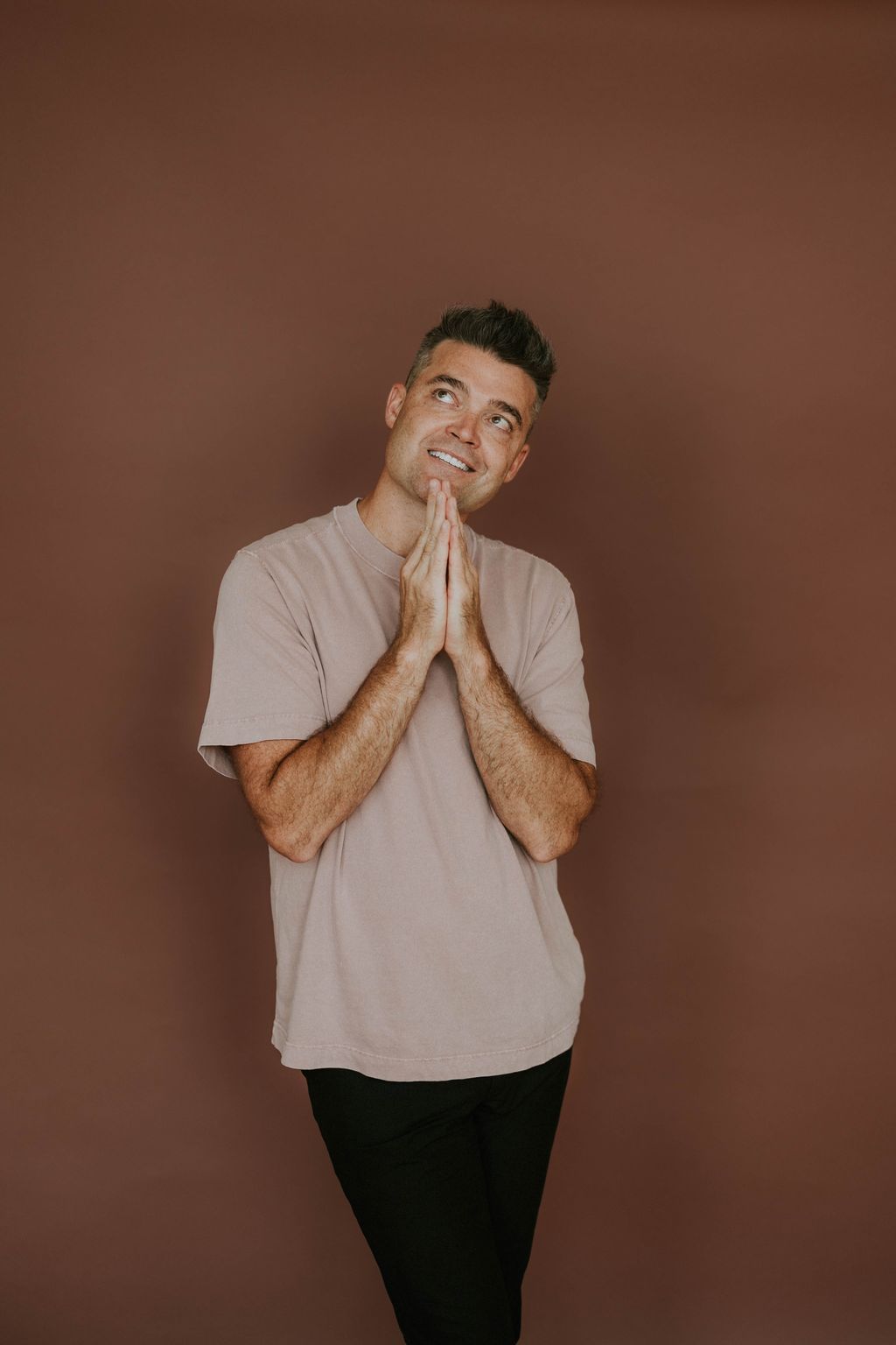 Nathan Morris smiling and looking up, hands pressed together, in a pink t-shirt against a warm brown wall.