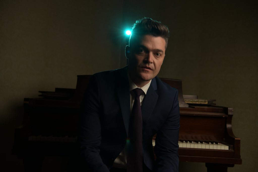 Nathan Morris in a suit playing piano under a teal stage light.