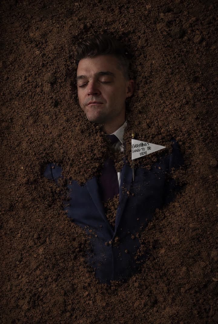 Overhead: Nathan Morris lies buried in dirt in a suit. A shovelful lands on him and he smirks at the camera.