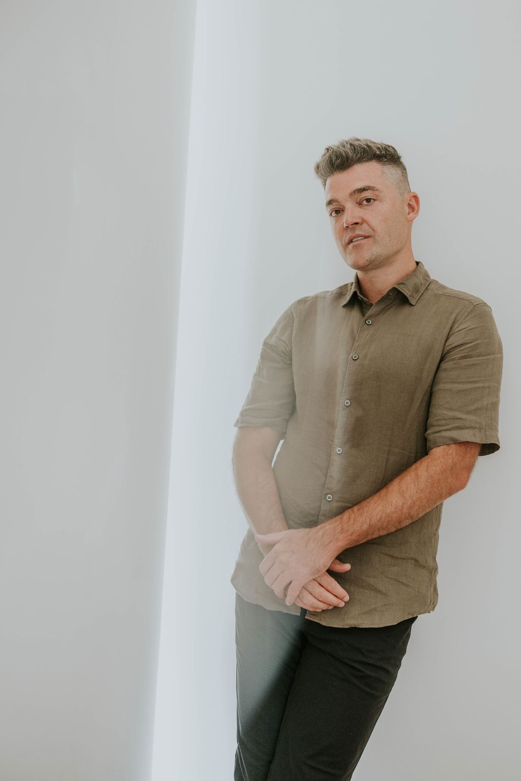 Nathan Morris in an olive linen shirt leaning on a white wall with his hands clasped.