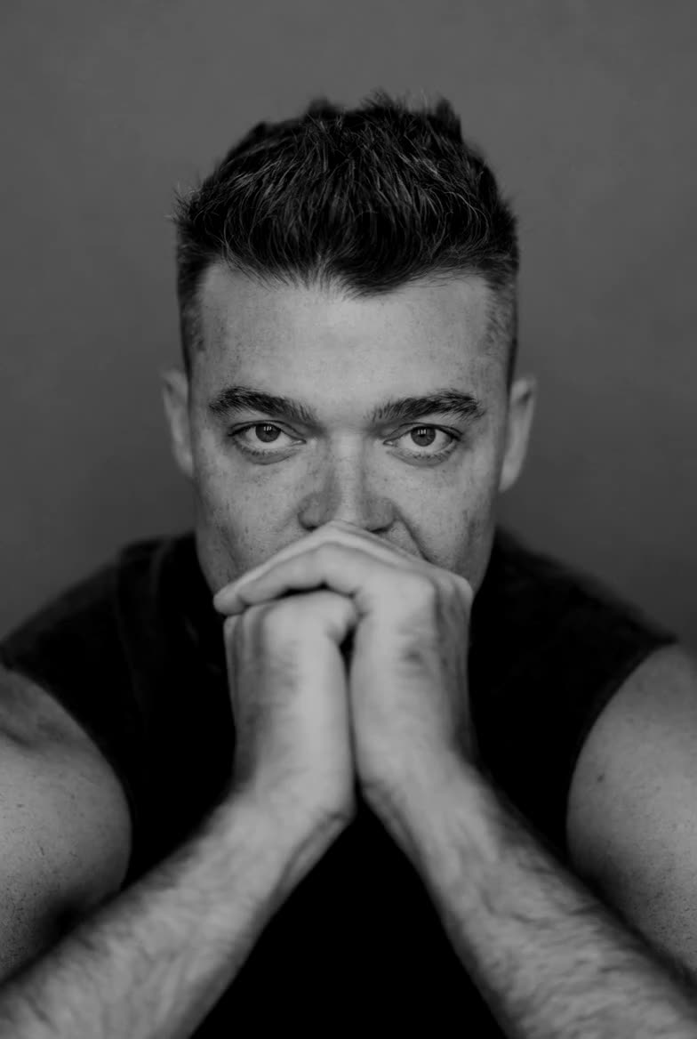 Nathan Morris, hands clasped at his mouth, holds your eye and smiles.